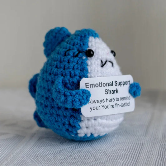 Crocheted emotional support shark toy holding a sign on a neutral background
