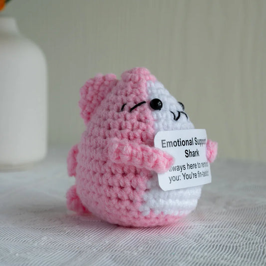Pink and white crocheted shark plush toy with a tag on a neutral background